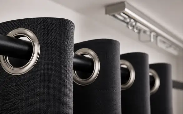 Close-up of eyelet ring curtains with brushed silver metal grommets threaded onto a matte black curtain rod, highlighting the heavy blackout fabric texture and curtain hardware.
